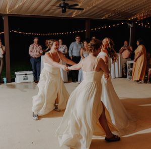 Two brides in white gowns dancing together at their outdoor wedding reception under string lights.