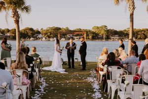 Outdoor waterfront wedding ceremony with palm trees and white chairs on grass.