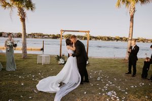 Romantic wedding kiss at waterfront ceremony with palm trees and scattered petals.