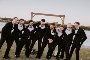 Groomsmen in black tuxedos pose playfully for group photo by the water.