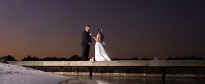 Wedding couple stands on wooden pier during golden hour with dramatic sunset sky.