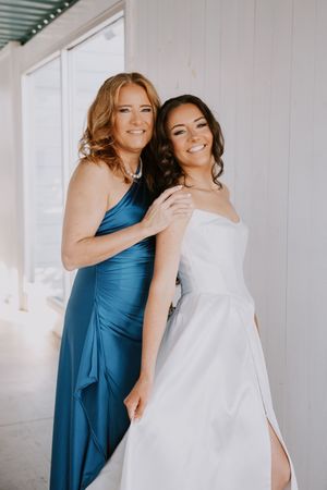 A bride in white and companion in teal blue dress share a joyful moment together during wedding preparations.