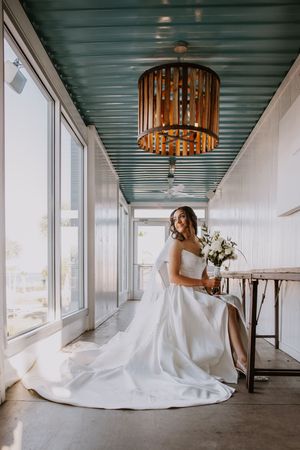 Elegant bride in white gown poses with bouquet near modern light fixture and windows.