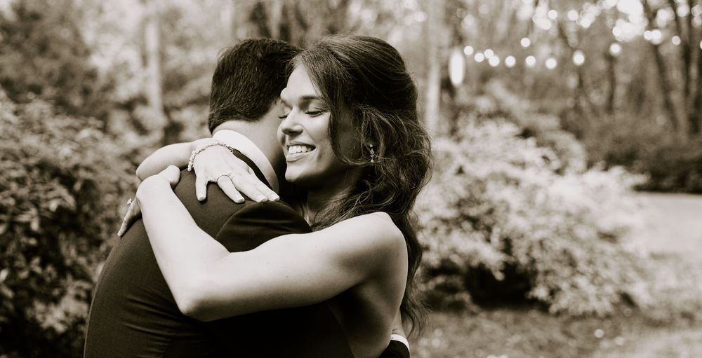 A joyful woman smiles while embracing a man outdoors in a lush garden setting, captured in black and white.