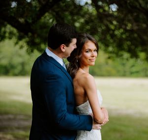 A bride in a white gown embraces her groom in a navy suit outdoors under lush green trees on their wedding day.