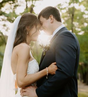 Bride and groom touching foreheads in golden sunlight, bride wearing veil and groom in navy suit outdoors.