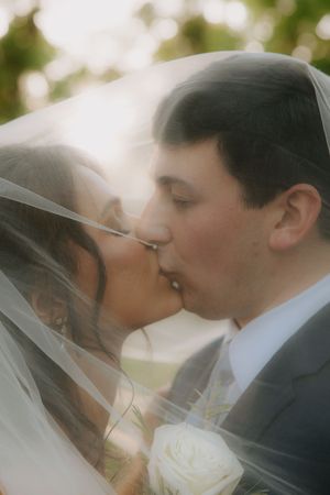 Bride and groom sharing a romantic kiss under a sheer wedding veil, with soft bokeh greenery in the background.