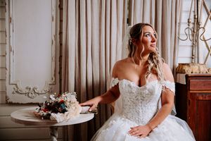Bride in off-shoulder lace ball gown sits by window with floral bouquet, gazing thoughtfully in elegant venue.