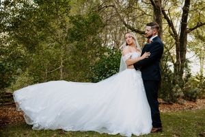 Bride in flowing white ball gown and groom in navy suit share romantic embrace outdoors surrounded by lush green trees.