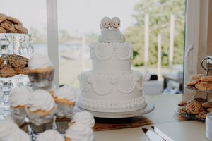 Three-tier white wedding cake with cute figurine toppers on a dessert table surrounded by cupcakes and cookies.
