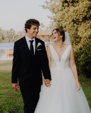 Happy newlyweds walk hand in hand outdoors, bride in white gown and groom in black suit, smiling joyfully.