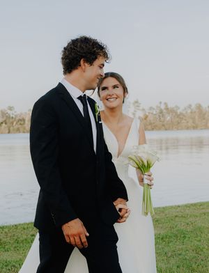 Bride and groom walking hand in hand outdoors near a lake, bride holding white calla lily bouquet in elegant wedding attire.