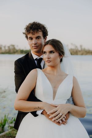 Bride and groom posing by a lake at sunset, bride in white gown with veil, groom in black suit.