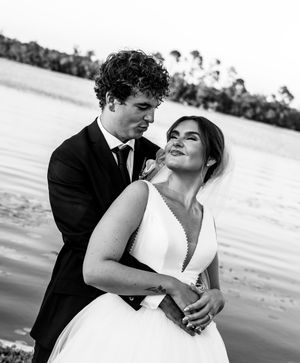 Black and white wedding photo of a couple embracing near a waterfront, bride in white gown with veil, groom in dark suit.