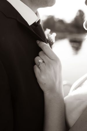 Black and white wedding photo of bride's hand with diamond ring grasping groom's suit lapel near a lake.