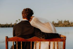 A newlywed couple in wedding attire sits on a wooden bench overlooking a serene lake surrounded by palm trees.