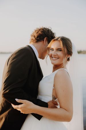 Smiling bride in white gown and veil embraced by groom in black suit during golden hour outdoor wedding.