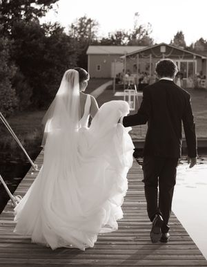 Bride and groom walking hand-in-hand on a wooden dock toward a lakeside wedding venue in a black and white photo.