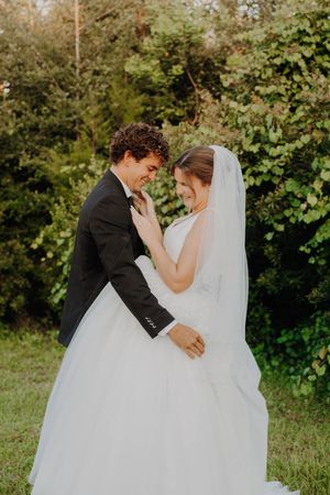 A bride and groom share a tender moment outdoors, smiling with foreheads together, surrounded by lush green trees.