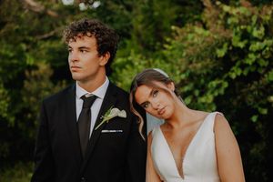 Elegant bride and groom in wedding attire posing outdoors with lush green foliage background.