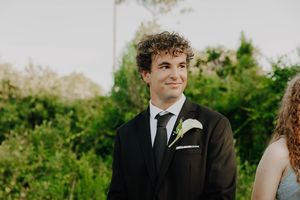 Young man in black suit with white boutonniere smiling outdoors at a wedding ceremony.