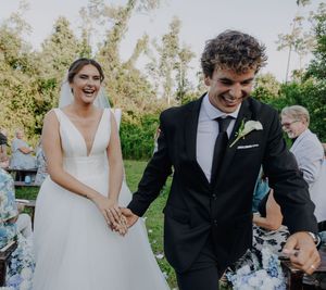 Joyful newlyweds in wedding attire holding hands and laughing during outdoor ceremony with guests in background.