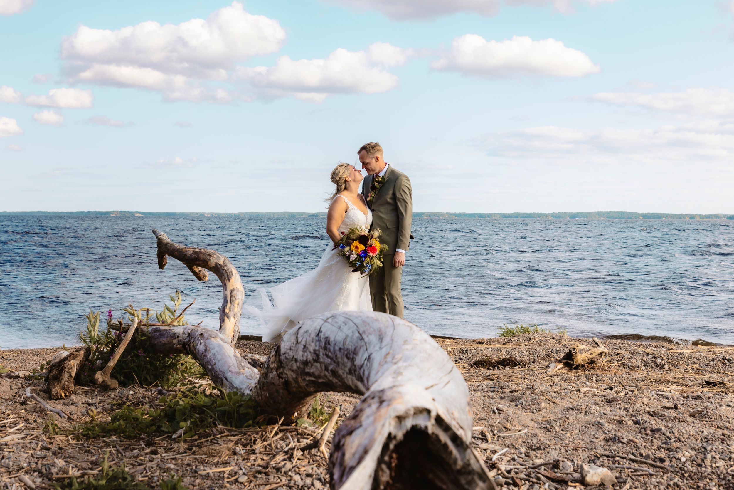 Brudpar kysser varandra på strand, med drivved i förgrunden och sjön Glan i bakgrunden.