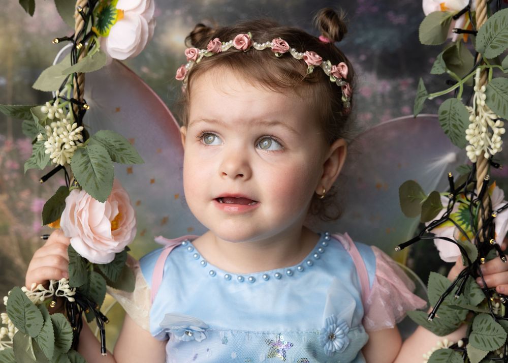 Dreamy portrait with floral crown and light blue dress surrounded by roses and white flowers against dark background.