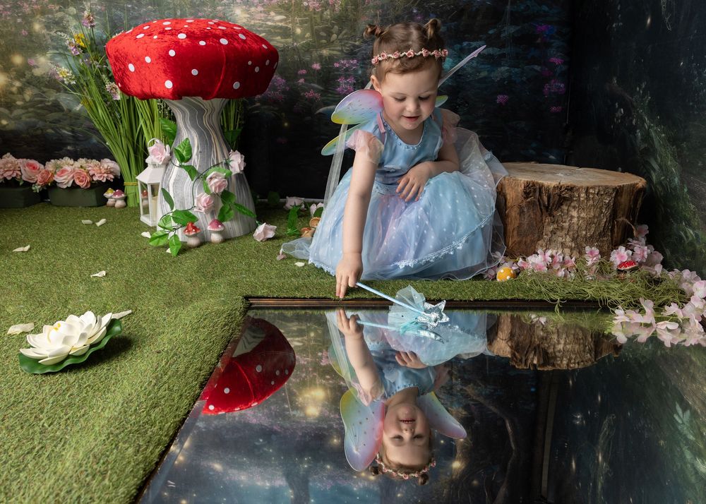 Whimsical garden scene with a red mushroom prop, tree stump, and mirror reflection on artificial grass with flowers.