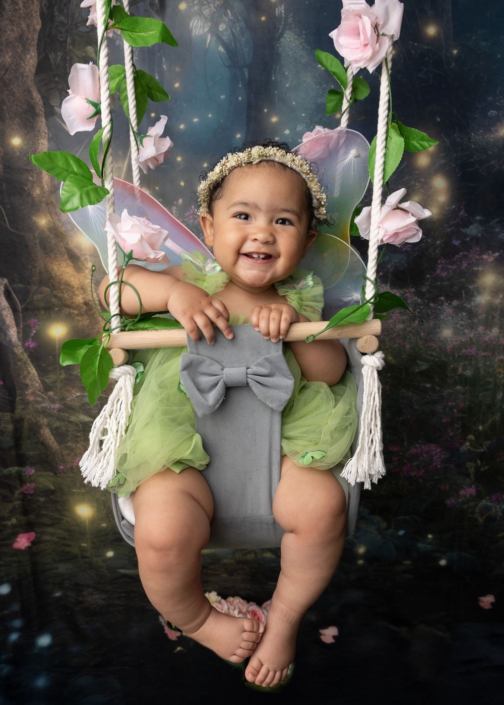 A baby wearing a green fairy outfit and flower crown sits on a decorated swing with pink roses and a magical backdrop.
