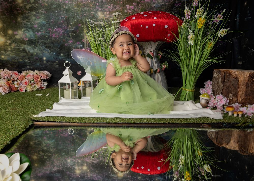 A child in a green fairy dress sits by a decorative garden scene with mushrooms, lanterns and reflective water.