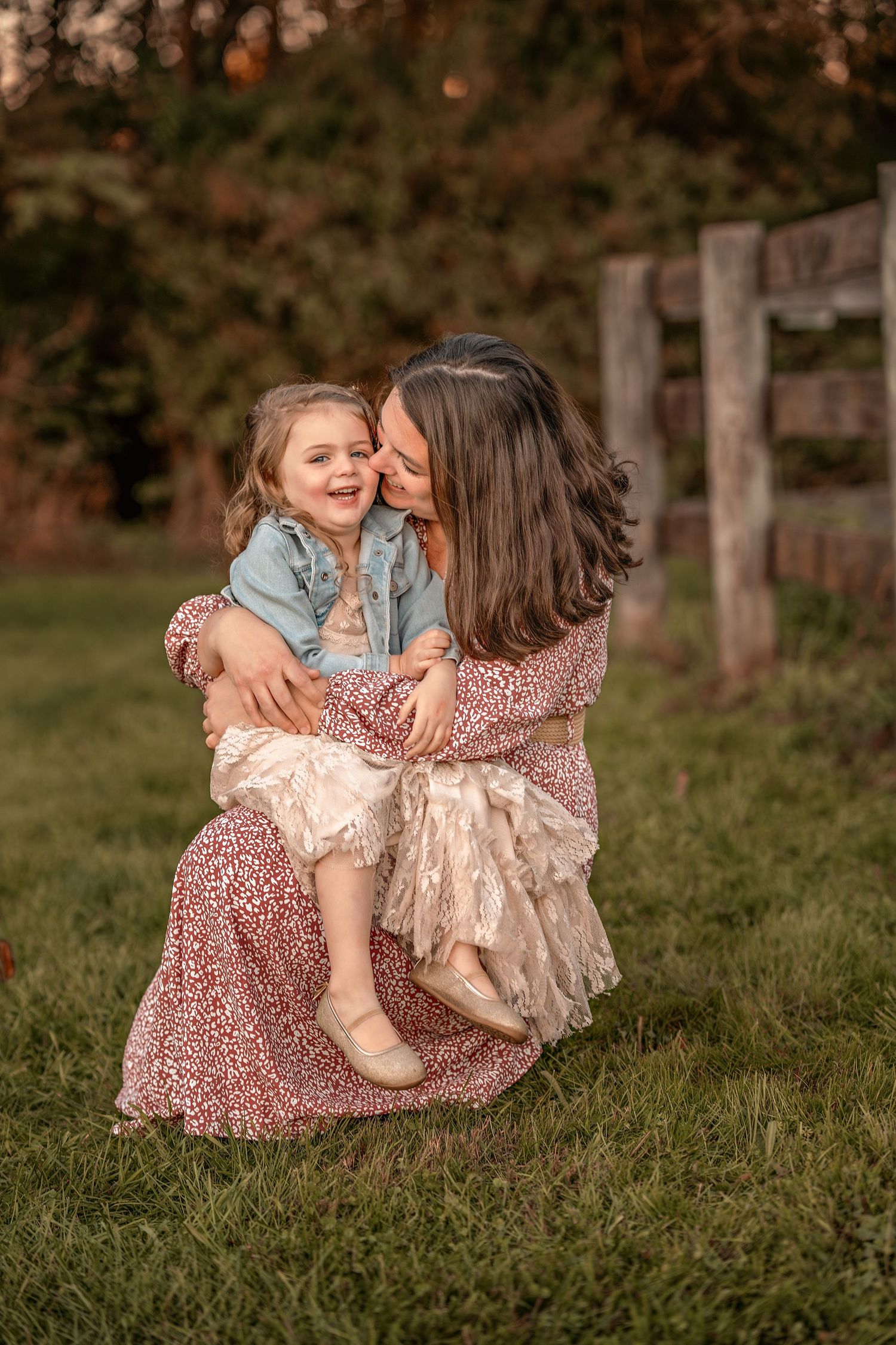 A tender moment shared between two people in floral dresses sitting on grass at sunset with a wooden fence in background.