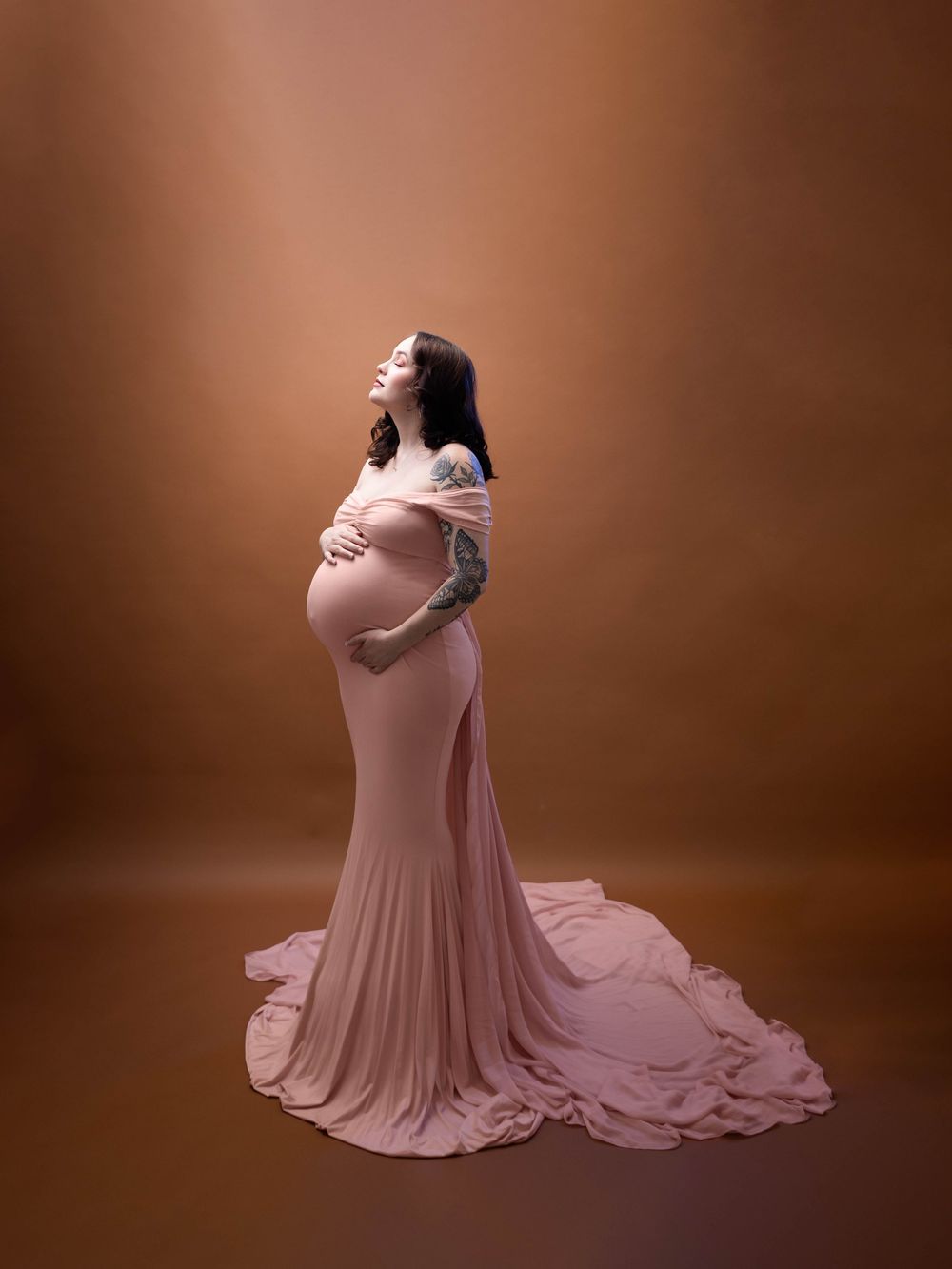 Maternity photo of a woman in a flowing blush pink gown posing elegantly against a brown backdrop.