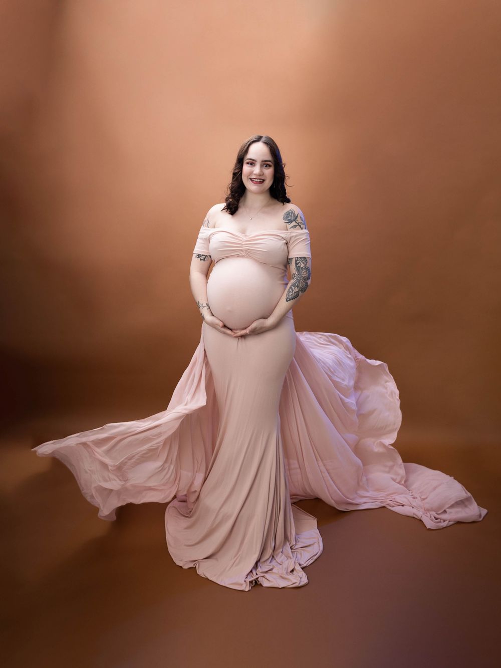 Expectant mother in blush pink off-shoulder gown poses against warm brown backdrop in maternity photo series.