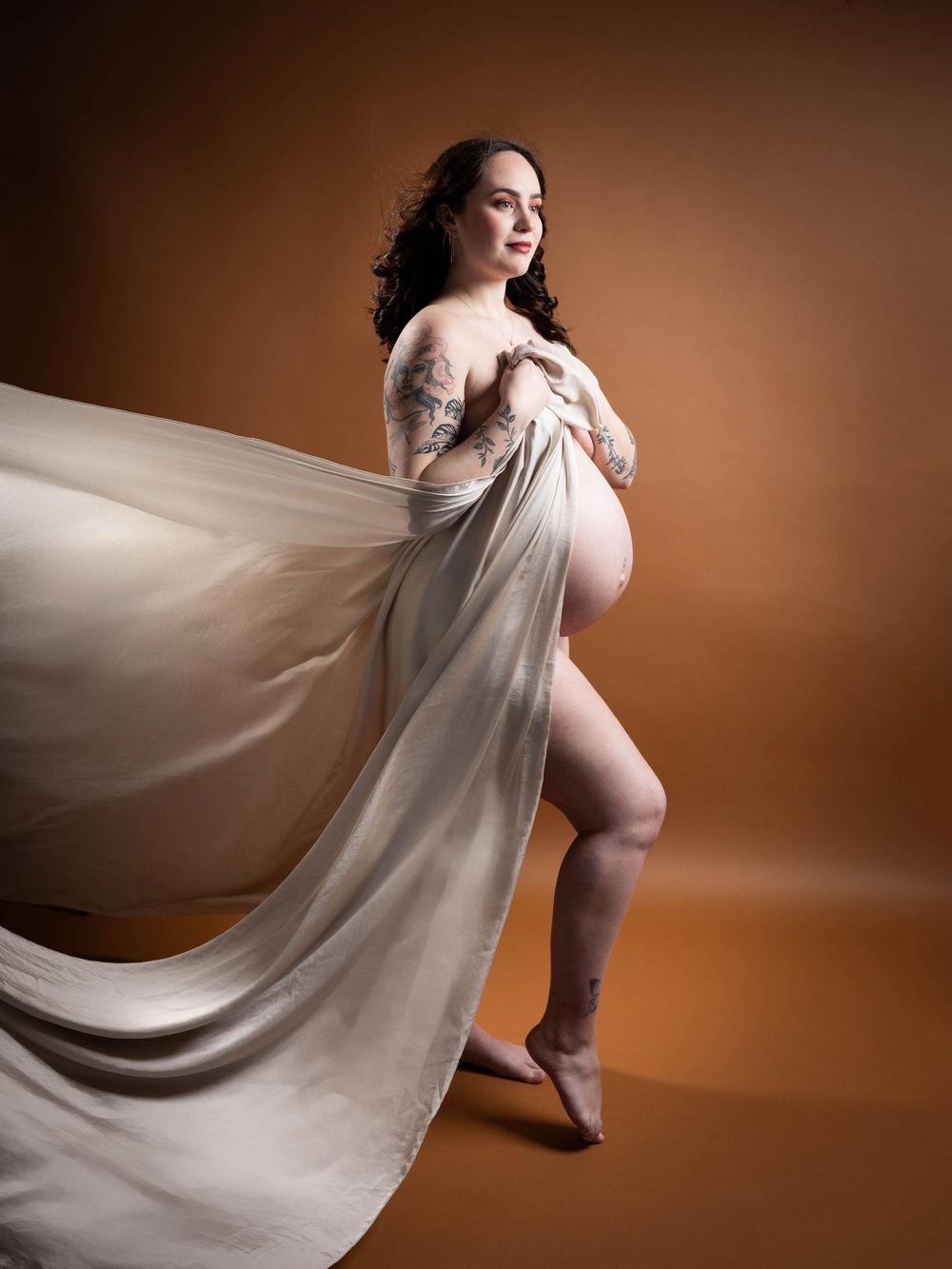 A flowing white sheer maternity gown creates ethereal movement against a warm brown studio backdrop.