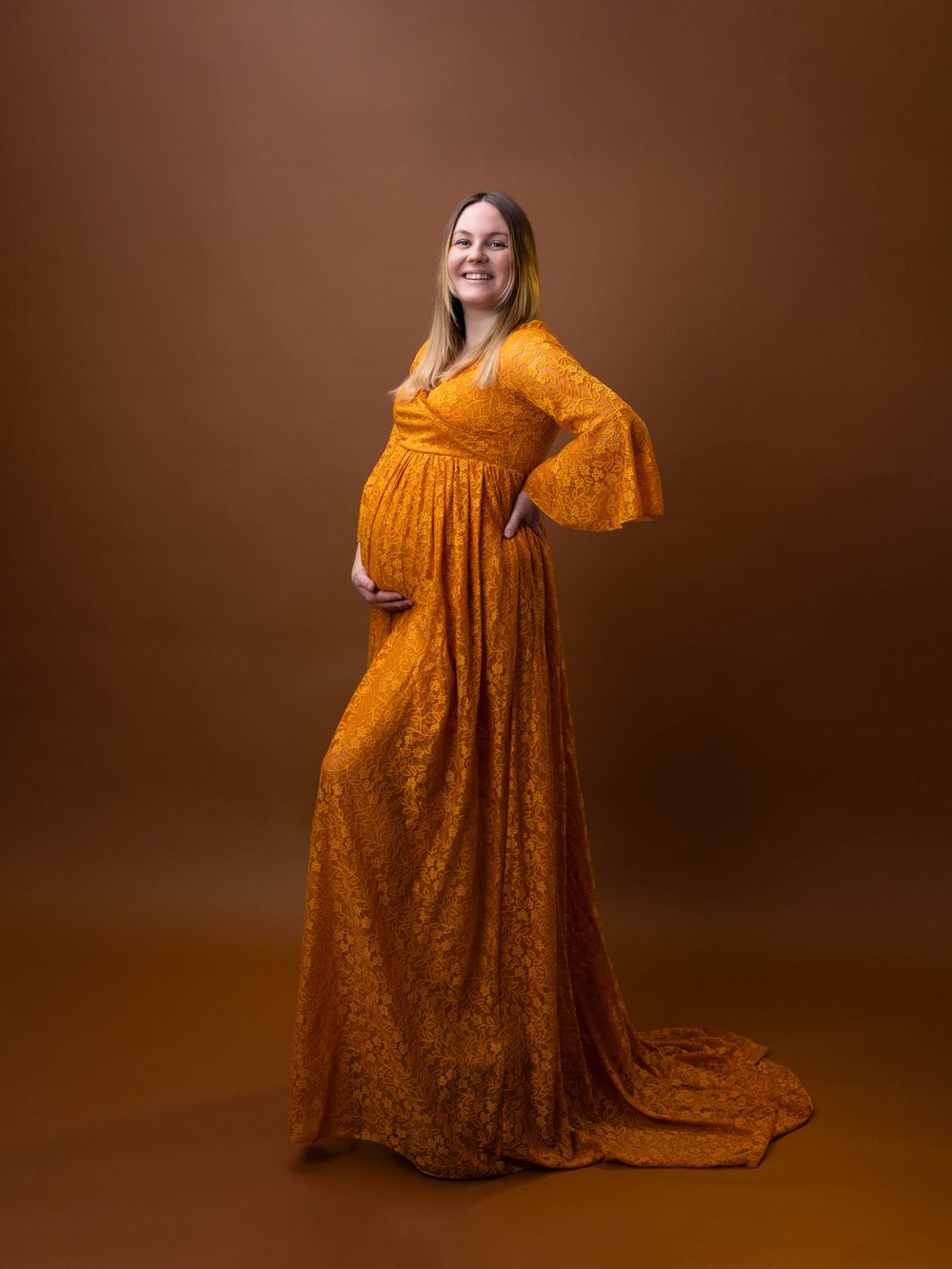 A woman in a flowing mustard yellow maxi dress poses gracefully against a brown studio backdrop.