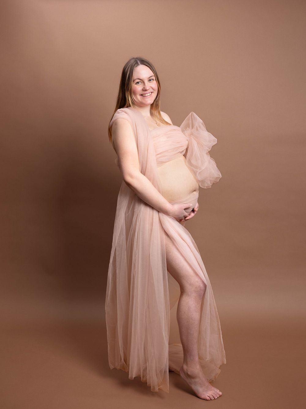 A person in a flowing peach tulle dress poses gracefully against a warm brown studio backdrop in a photo series.