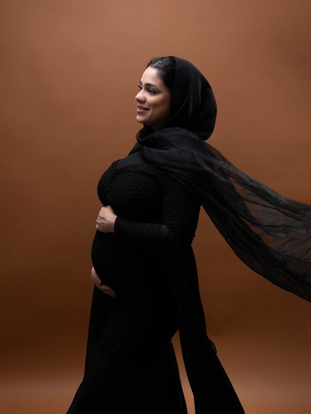 A pregnant person in a flowing black dress and hijab poses against a brown backdrop with graceful movement.