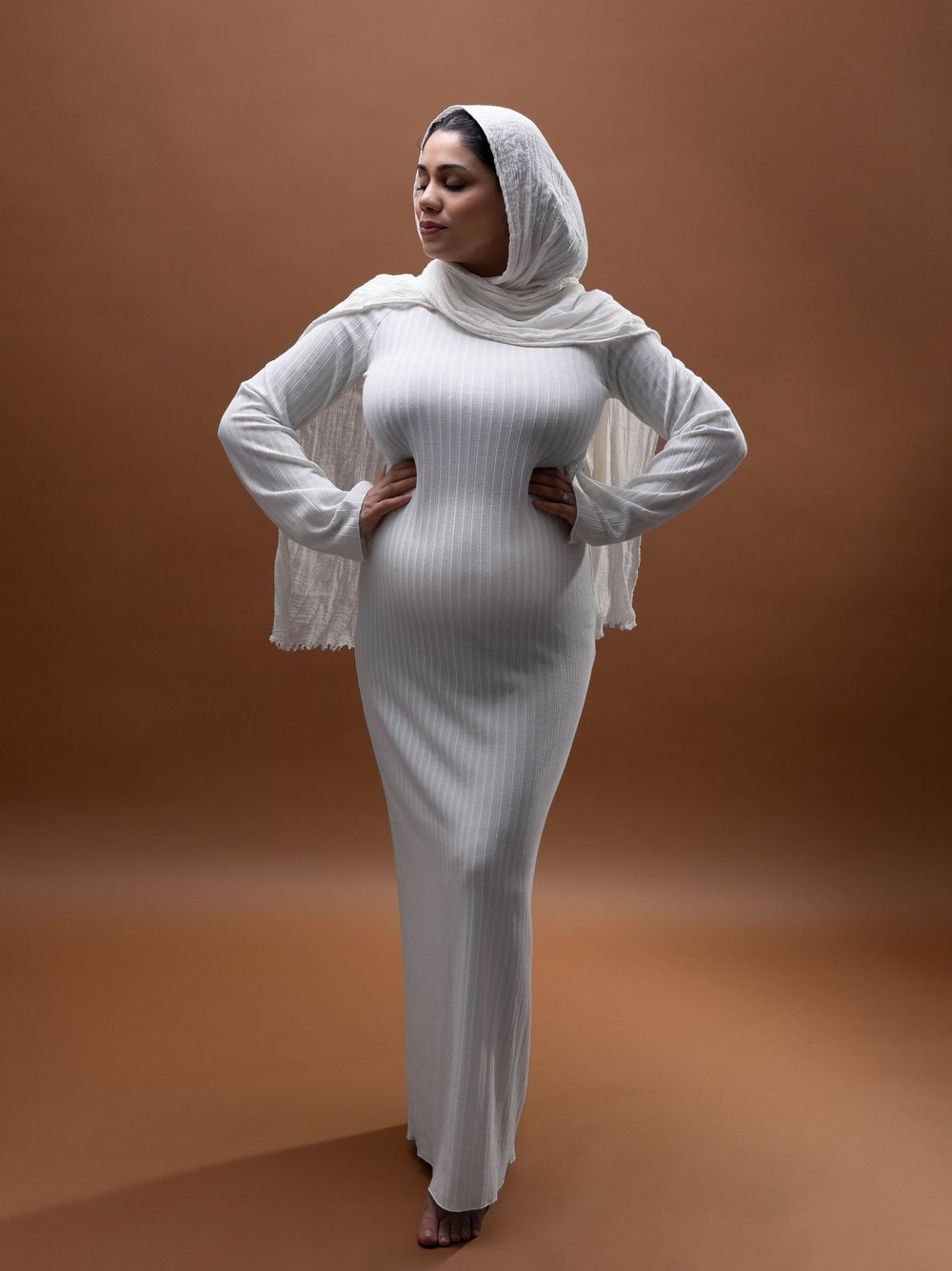 A figure in a white ribbed bodycon dress and matching hijab poses confidently against a brown studio backdrop.