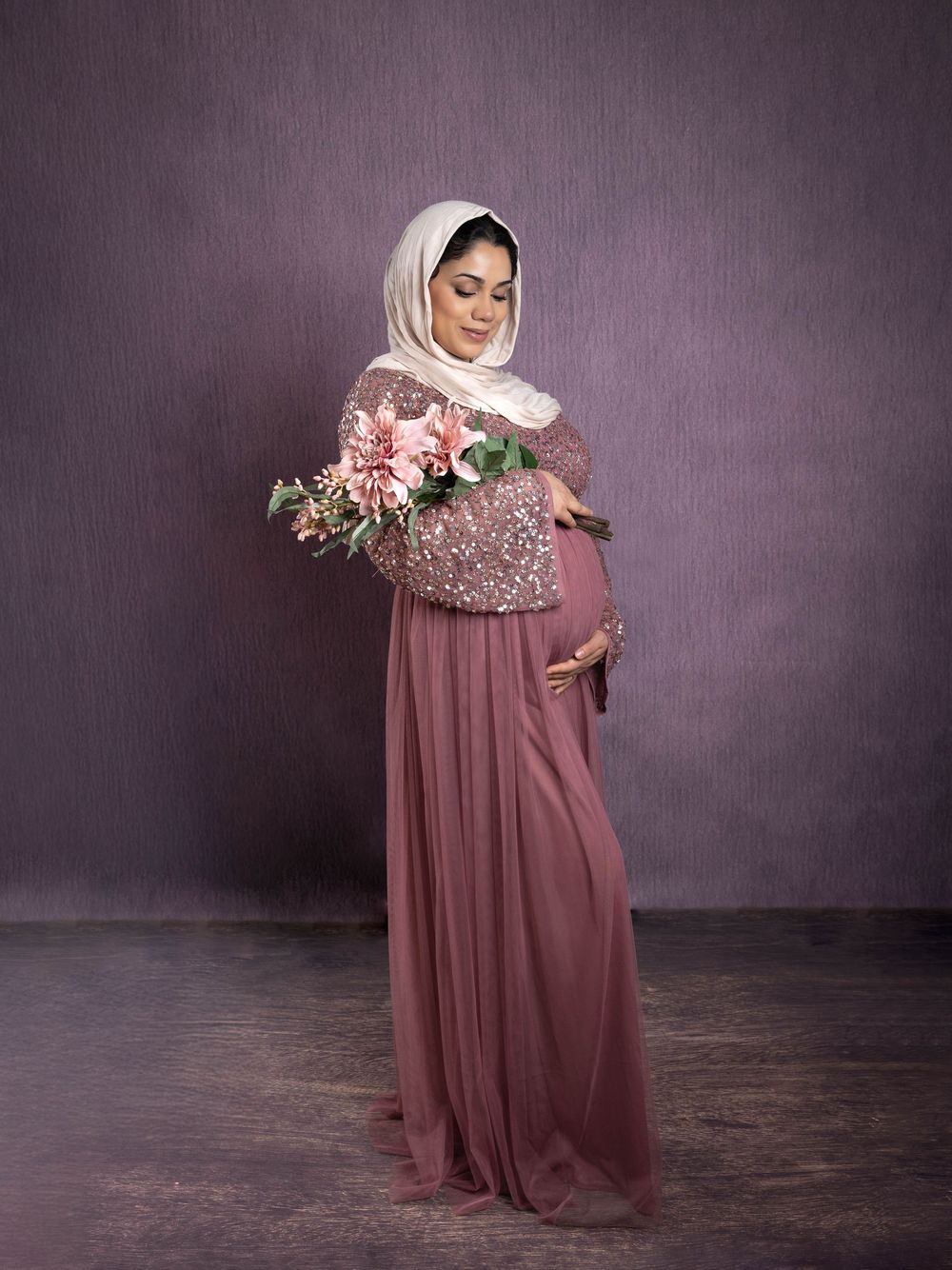 Model in flowing mauve gown poses gracefully against dark backdrop in elegant maternity photoshoot series.