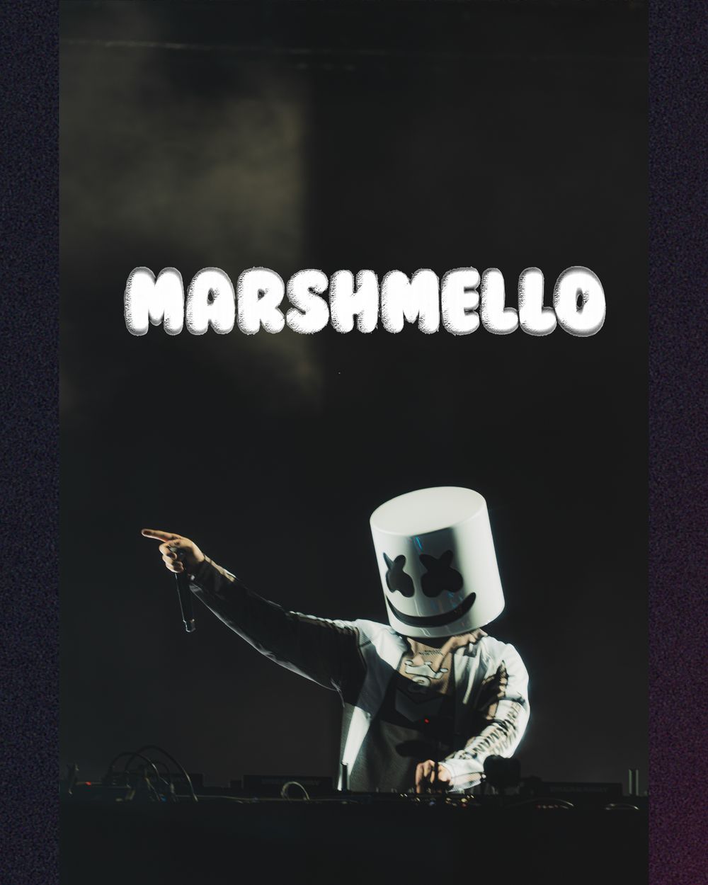 Marshmello DJ performing live on stage wearing iconic white helmet costume with DJ equipment in dark concert setting.