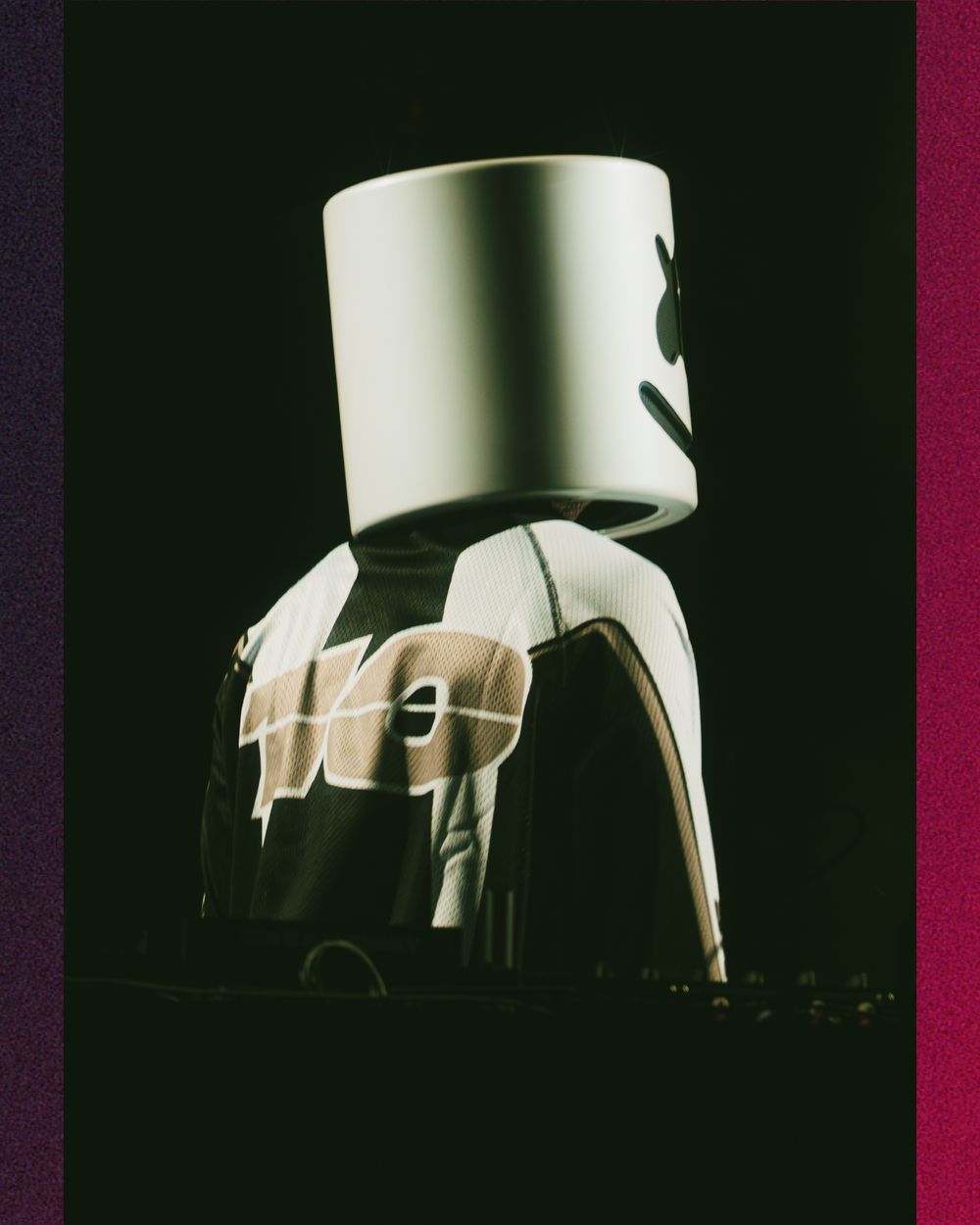 DJ Marshmello performing on stage wearing iconic white helmet mask and black jersey under dramatic dark lighting.