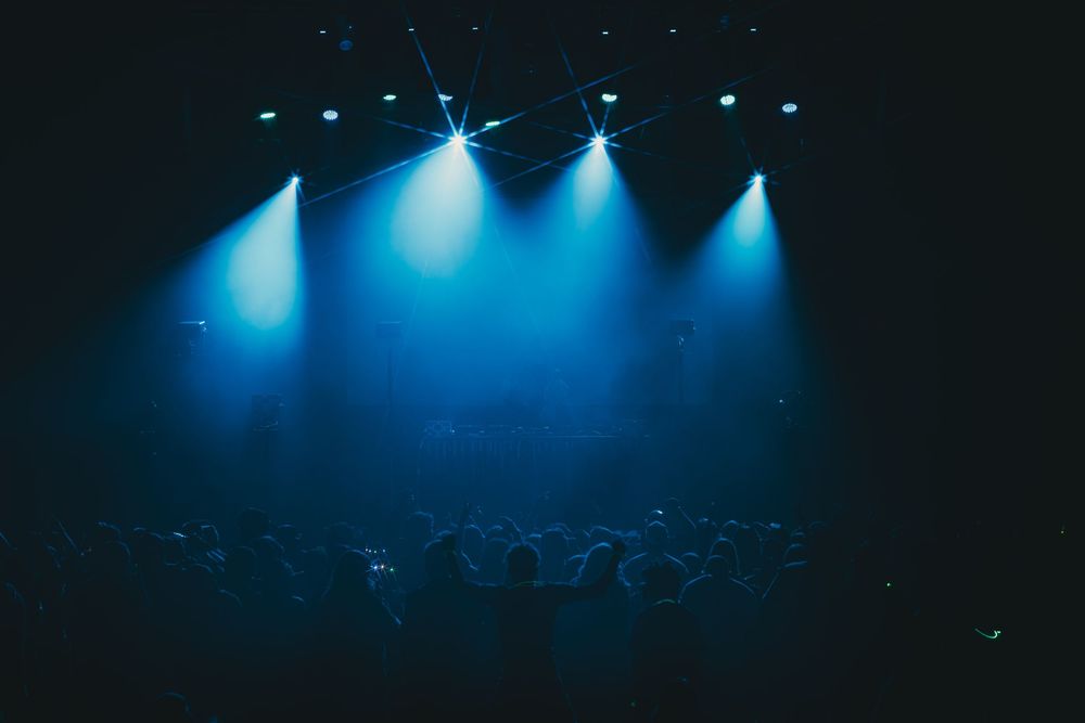 Blue stage lights illuminate a dark concert venue filled with silhouetted crowd members at a live music event.
