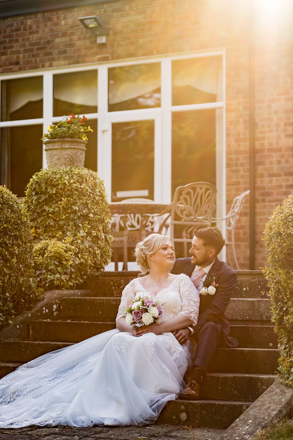 Newlywed couple sits together on stone steps at sunset with bride holding bouquet in lace gown.