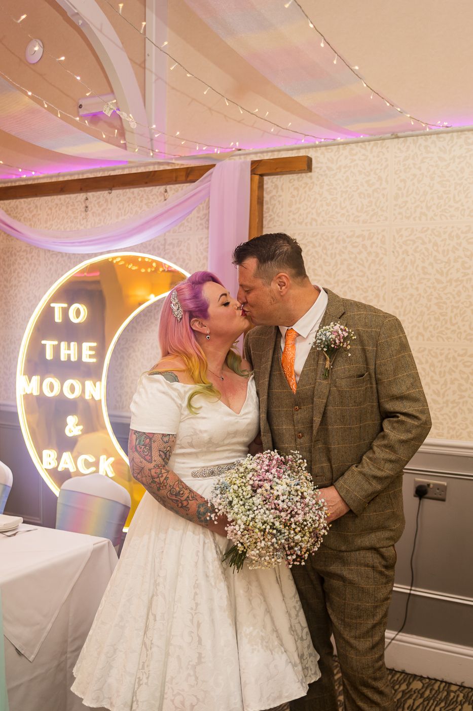 Wedding couple shares intimate moments in a reception venue with illuminated neon signs and soft pink lighting.
