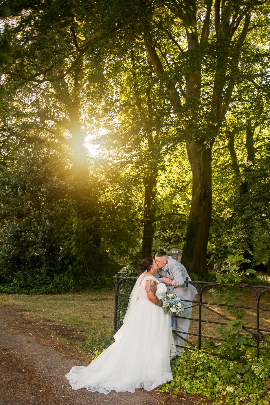 Sunlit forest scene with a couple in wedding attire embracing near a rustic fence during golden hour.