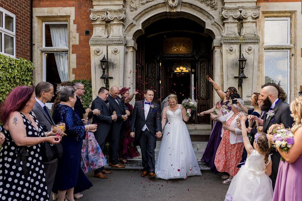 Wedding guests gather outside a historic brick building with arched doorways during a celebration.