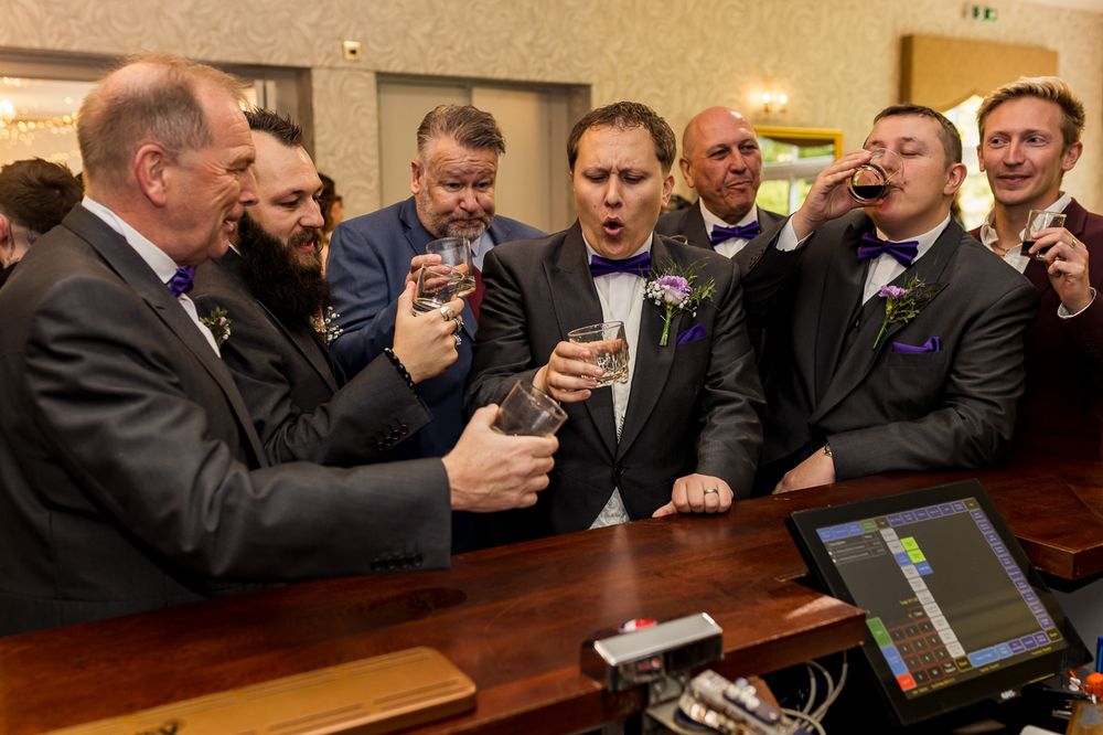 A group of people in business attire gather around a bar counter while holding drinks and socializing.