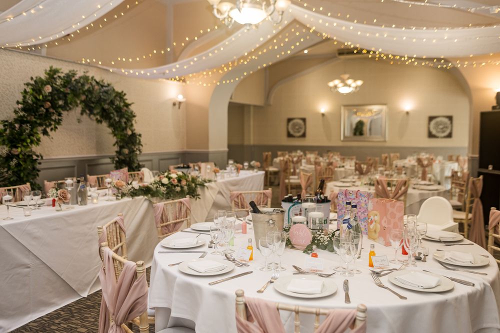 Elegant wedding reception venue with round tables, white linens, pink accents, and twinkling string lights overhead.