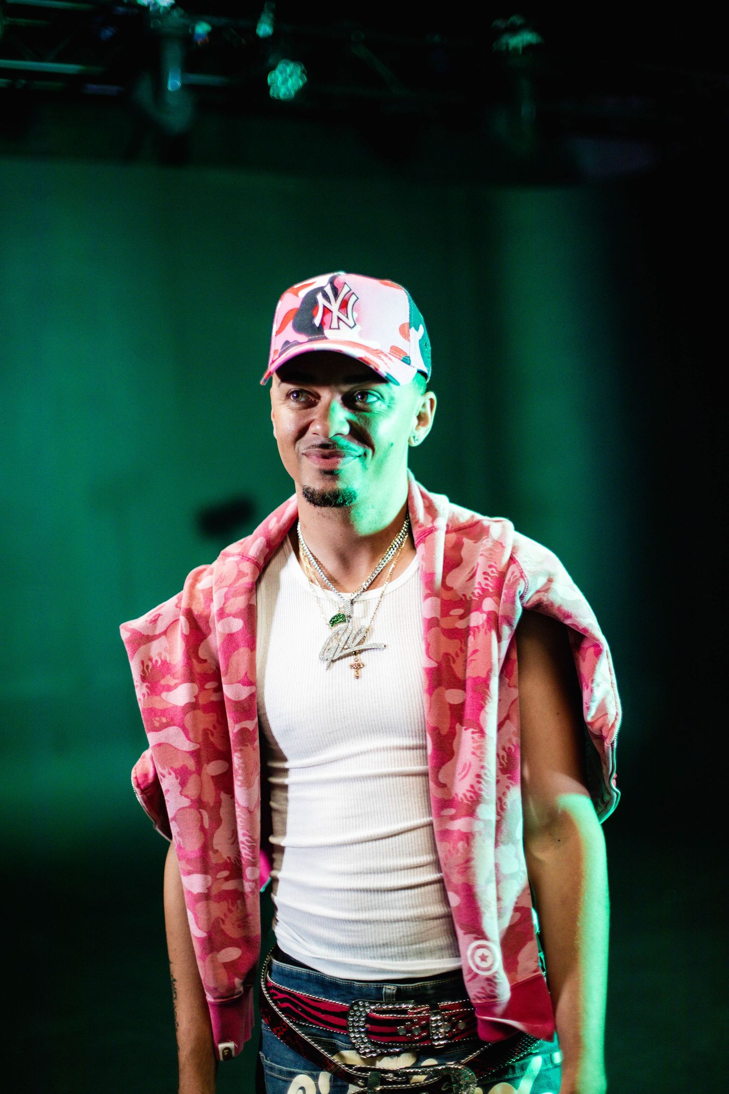 A person wearing a white tank top and pink floral vest poses against a green background.